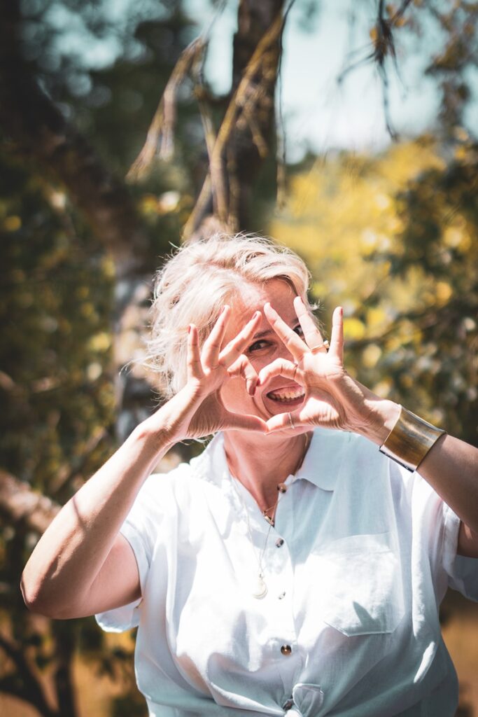 Frau lächelt in der Natur und formt mit den Händen ein Herzsymbol – Sinnbild für Lebensfreude, Selbstverbindung und den Ruf des Lebens.