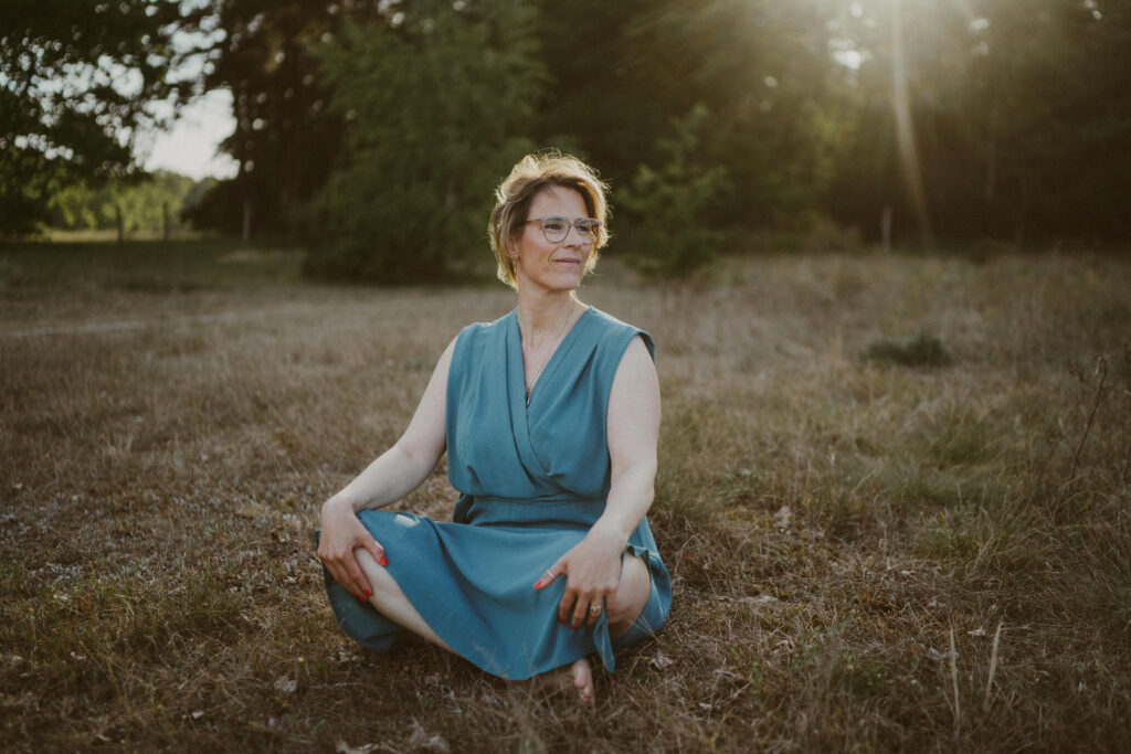„Frau in blauem Kleid sitzt barfuß und entspannt im Gras, Sonnenlicht fällt durch die Bäume – Symbol für Achtsamkeit, Naturverbundenheit und innere Balance.