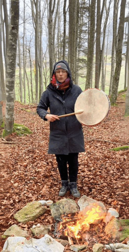 Frau mit Trommel steht im Wald an einem Feuer – Symbol für Rituale in der Natur, Achtsamkeit und spirituelle Verbindung.