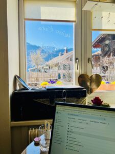 Arbeitsplatz in der Lebensinsel Brienz: Laptop auf dem Tisch, Blick durch das Fenster auf Berge, Häuser und Frühlingsblumen – Sinnbild für Remote-Work in inspirierender Umgebung. Meine Selbstständigkeit aufbauen war hier angesagt