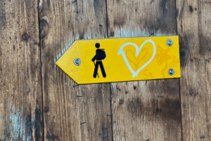Gelber Wegweiser mit Wander-Symbol und Herz auf Holz – Symbolbild für den Ruf des Lebens und das Finden der eigenen Lebensmission.