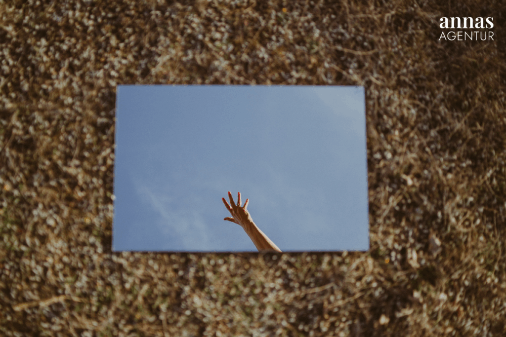 Spiegel auf dem Boden, in dem sich ein klarer Himmel zeigt, während eine Hand nach oben ragt – Symbol für Selbstreflexion, Perspektivwechsel und innere Ausrichtung.