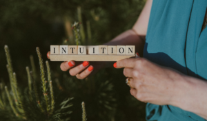 Frau hält das Wort „Intuition“ aus Holzbuchstaben in den Händen, umgeben von Natur – Symbol für innere Wahrnehmung und verkörpertes Wissen