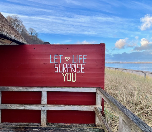 Rote Strandhütte an der Küste mit der Aufschrift „Let life surprise you“ – ein Ort der Pause und Ausrichtung, der für Offenheit gegenüber inneren Signalen steht.